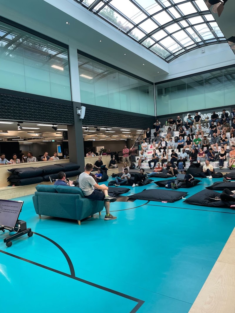 A photo taken from the front of the Roo pitch, looking up at the bleachers which are almost full with attendees. Some atttendees are sitting around the side of the pitch on on the benches as well as some on the laying mats. Dan and Daniel can be seen on the sofa answering questions
